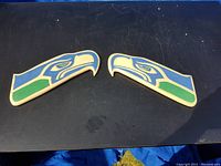 Pair of flat wooden carvings depicting seahawk heads painted blue, green, and white, laid out side-by-side showing both pieces