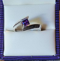 Sterling silver ring with square cut amethyst stone set asymmetrically in a white cushioned box with purple-striped lid.