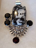 Photo of a large sterling silver pendant with an intricate design, featuring a central black and white rectangular stone, three faceted black stones, and one garnet stone.