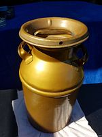 Gold-painted vintage metal milk can with wide neck and handles shown from side angle in daylight against blue background.