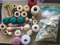 Crochet supplies including many thread and yarn spools and balls in various colors, some metal crochet hooks, and a large zip-lock bag of crocheted items shaped like pineapples.