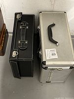 Top view of two cases; one black with leather-like texture and metal clasps, the other silver aluminum with black handle and clasps.