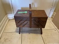 Full front view of the closed vintage wooden accordion sewing basket with stand on tile floor.