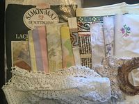 Assorted folded fabric pieces and lace trims including embroidered white linens, pastel and floral prints, and crocheted lace doilies.
