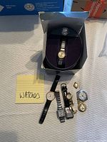 Photo showing six watches and one pendant watch, with a blue and black box containing a large round silver-tone wrist watch, and other watches laid out on fabric surface.