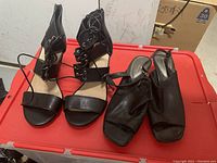 Black lace-up open-toe heels and black leather open-back square-toe shoes shown together on red container