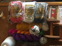 Photo showing packaged Phentex yarn skeins in red, white, and black along with assorted colored yarn skeins in various textures and colors on a wooden surface. Includes white textured yarn balls and a pink-purple skein.