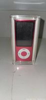 Front view of red Apple iPod Nano inside its clear plastic case showing screen and white click wheel, brand new.