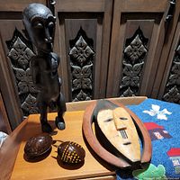 Front view of male wooden African statue, handmade coconut shell turtle and bug figurines, and hand-carved painted wooden mask on wooden surface.