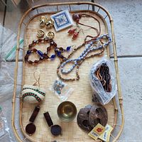 Top view of assorted loose jewelry and accessories laid out on woven tray including beaded necklaces, clip-on earrings, carved pendant, small beads, wooden pendants, a brass bowl, and decorative tile piece.