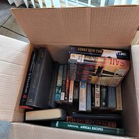 Top-down view of an open cardboard box filled with various paperback and hardcover books, including novels and cookbooks.