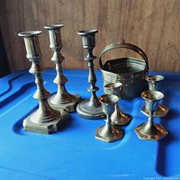 Group photo showing three tall brass candlestick holders, three smaller brass candle holders, and one brass basket with handle, all placed on a blue surface against a wooden background.