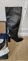 Black leather knee-high boot standing upright beside its box showing the high block heel and almond toe.
