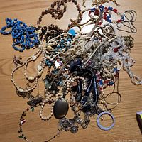 Mixed pile of tangled costume jewelry pieces including beaded necklaces, chain necklaces, bracelets, and watches on a wooden surface.
