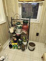 Photo of a black metal decorative indoor plant stand holding multiple various indoor flower pots including ceramic, plastic, and terracotta types. Additional potting sprays, seed packets and planted items are placed on and around the stand.
