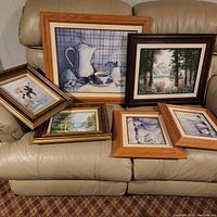 Front view of six framed art prints arranged on a beige leather couch showing various scenes including still life and landscapes