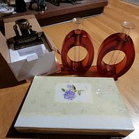 Two thick red resin flower stem vases, photo album with floral cover, and boxed Eoros candle holder figurine on floor.