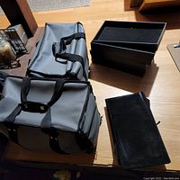 Two gray nylon jewelry display bags on a wooden floor with 6 black felt-lined trays and several black jewelry display plaques laid out beside them.