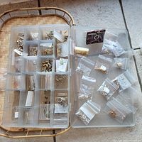 Overview of plastic storage case with multiple compartments filled with gold and silver jump rings, head pins, and small jewelry components in plastic bags.