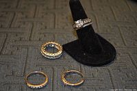 Four silver rings on cloth surface with one ring displayed on a black stand featuring a large square CZ stone center and smaller CZ stones on band.