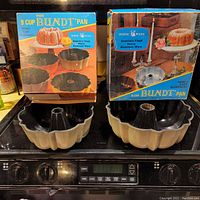Two vintage Nordic Ware bundt cake pans, one 9-cup and one 12-cup, shown with their original colorful boxed packaging on a stovetop.