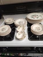 Full view of all china pieces arranged on stovetop showing multiple stack of plates, bowls, cups, and saucers with the pink rose pattern and detailed gold lattice rim.