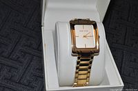 Gold-tone Anne Klein wristwatch on white pillow inside open Anne Klein box on dark fabric.