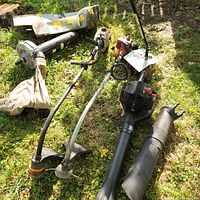 Lot of yard tools displayed on grass including gas-powered weed wackers and blower with collection bag attachment.