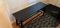 Black wooden bench positioned against a wall on a patterned rug near the entrance-way mat and shoes.