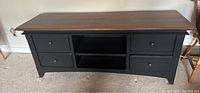 Full front image of the console table showing four drawers with black knobs, two open center shelves, natural wood top, black base and legs.