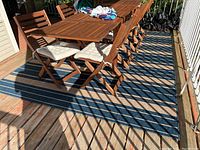 Photo shows the striped blue and beige indoor/outdoor rug placed on a wooden deck beneath a wooden table and chairs. The rug size measures approximately 8 by 10 feet as described.