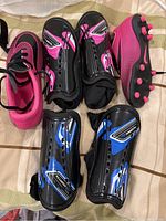 Photo showing a pair of pink and black kids soccer cleats alongside two pairs of shin pads, one pink and black, one blue and black, all with adjustable straps.