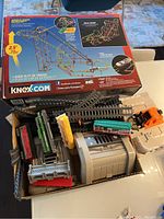 Photo shows K'NEX roller coaster set box on top with the HO scale train components and track pieces laid out below. Several train cars in multiple colors visible along with gray tunnel and track bridges.