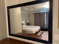 Photo showing large rectangular mirror with black frame mounted on a wood-paneled wall, reflecting bedroom interior.