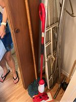 Photo of a red and black mop with blue mop head leaning against a wooden cabinet and an ironing board behind it.