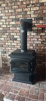 Black metal natural gas stove resembling a traditional wood-burning stove against a brick wall. Features glass door and vent pipe.