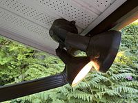 Dual-head outdoor floodlight mounted under eave with one lamp lit, showing condition and design featuring a motion and light sensor above the two black cone-shaped lamp heads.