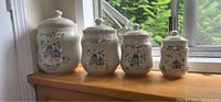 Four Japanese stoneware kitchen jars arranged in size order on wooden surface near window, decorated with house and floral motifs.