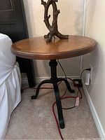 Round table with smooth solid wood top, supported by black metal tripod legs, shown next to bed.