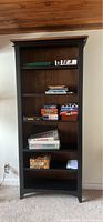Photo of a tall black solid wood bookshelf with several shelves, some holding books and board games. The bookshelf is against a wall with a wooden ceiling.