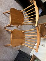 Photo shows two wooden chairs with spindle backs, straight legs, and a natural wood finish on a carpeted floor in indoor setting.