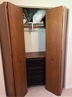 Open double wood bifold closet doors revealing a closet interior with shelves and dark drawers inside, showing construction and hardware details.