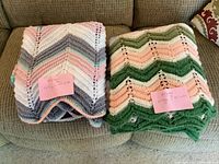 Two folded knitted afghans on a sofa with size notes attached. One afghan has pastel pink, gray, white, and mint green chevron stripes; the other has green, peach, white chevron stripes.