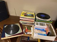 Table with stacked vinyl records, some out of sleeves, magazines, and box with VHS tapes visible.