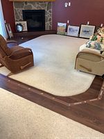 Area rug shown in a living room with hardwood floor borders, sofa, recliners and fireplace in the background. Rug is off-white and thick plush.