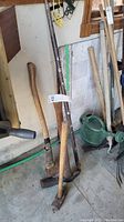 Photo showing five long-handled tools including axes, sledgehammer, pickaxe, and pry bar leaning against a garage wall.