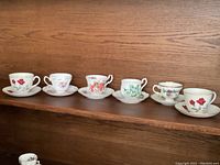 Six different sets of English china teacups and saucers each with floral patterns on a wooden shelf.