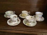 Photo showing five English fine china teacup and saucer sets with varied floral patterns and gold trim on a dark wood shelf.