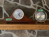 Both clocks displayed on a wooden shelf in front of a stone wall, showing size and style contrast.