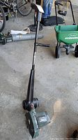 Full view of the Black & Decker garden edger trimmer standing upright on a concrete garage floor near other garden tools.
